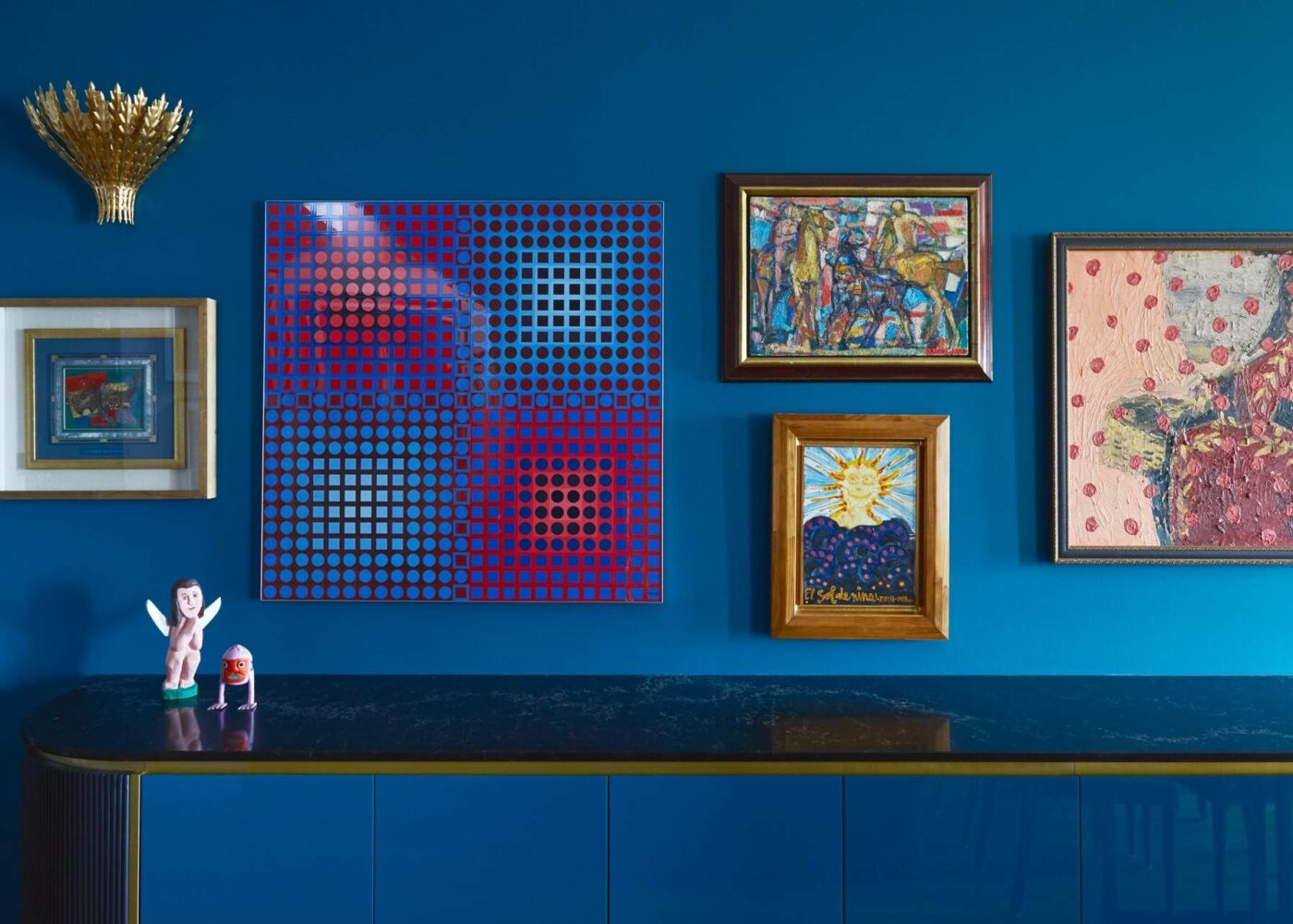 A modern interior showcasing a blue wall adorned with various artworks, including a prominent dotted pattern piece in red and blue, framed pictures, and decorative figurines.