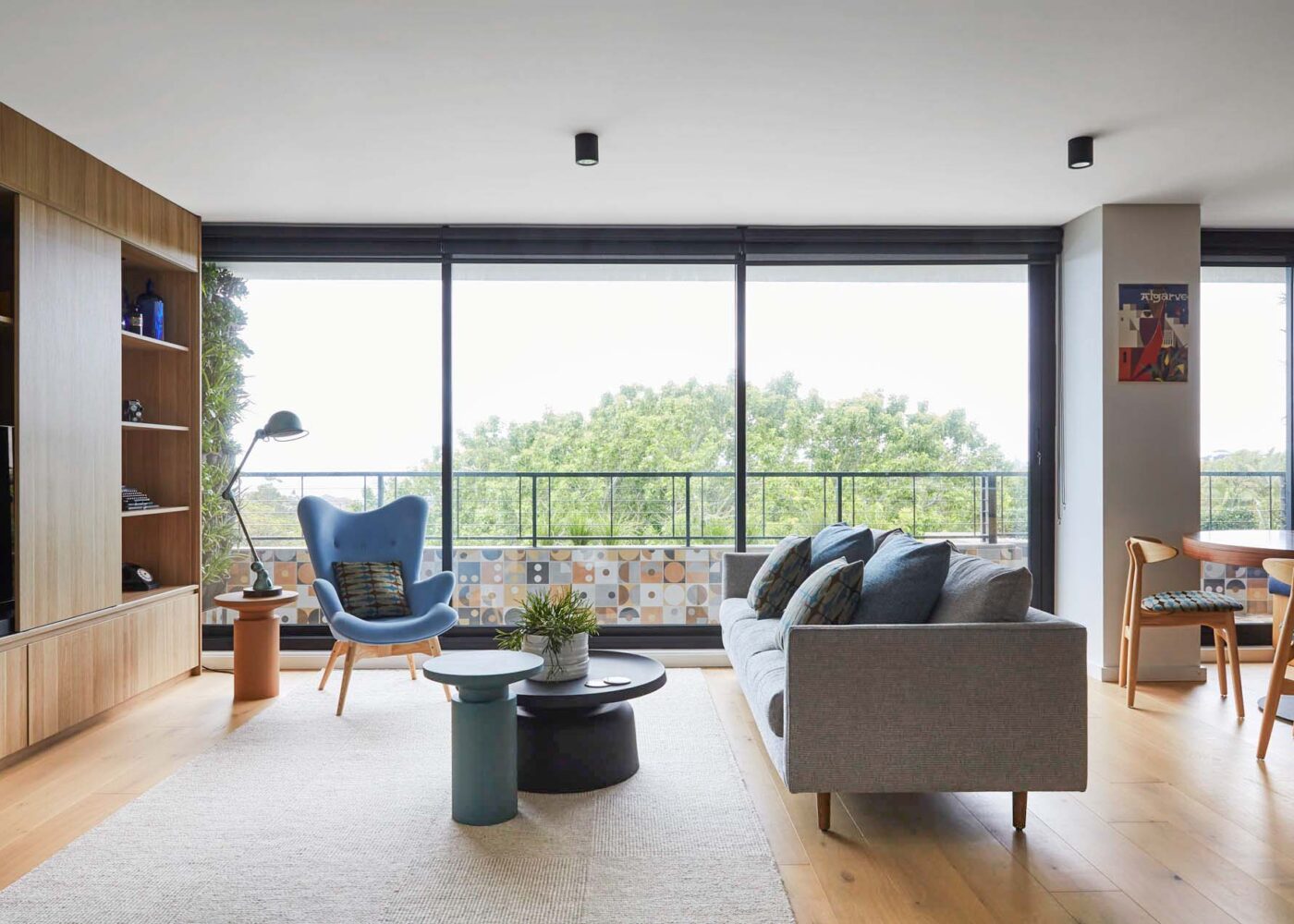 A modern living room featuring large windows with greenery outside, a stylish blue chair, a cozy gray sofa, and contemporary furniture pieces. Ideal for interior design inspiration.